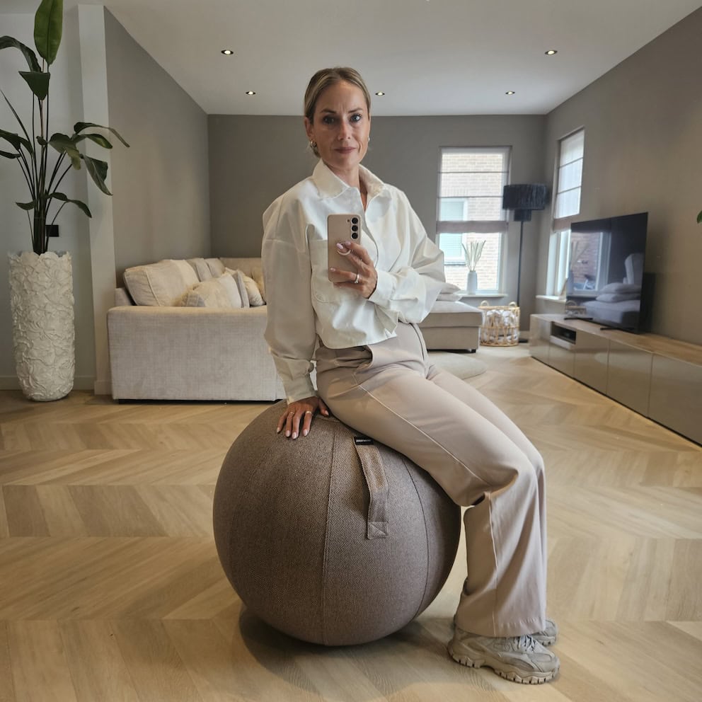 Elegante vrouw in beige en witte kledij zittend op een fitnessbal in een modern woonkamer met houten vloer en grote ramen. Moderne interieurstijl, comfortabele zithoek, lichte kleuren, planten en televisie voor een gezellige sfeer.