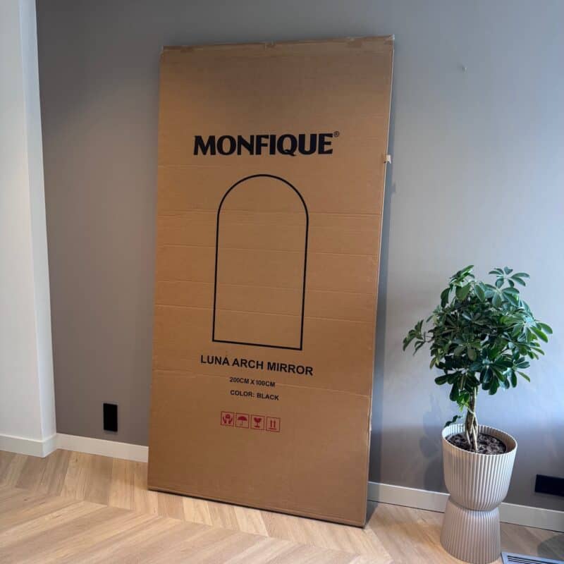 Grote kartonnen doos voor een Luna boogspiegel van Monfique.nl, geplaatst in een modern interieur met houten vloer en een groene plant in een witte pot.