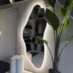 Unieke spiegel met indirect verlichting in moderne woonomgeving voor stijlvolle garderobe of interieuraccent. Spiegel met organische vorm en LED-verlichting, inclusief opbergkast en een grote groene plant voor een trendy interieur.