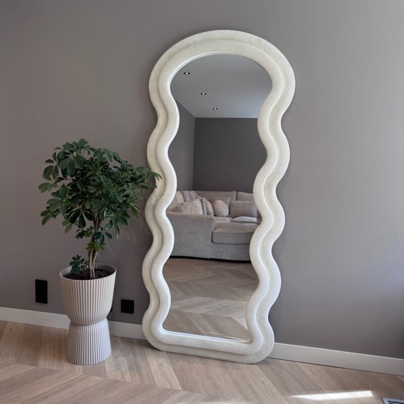 Elegante witte spiegel met golvend frame in een modern interieur met grote plant en comfortabele woonkamer in de achtergrond.